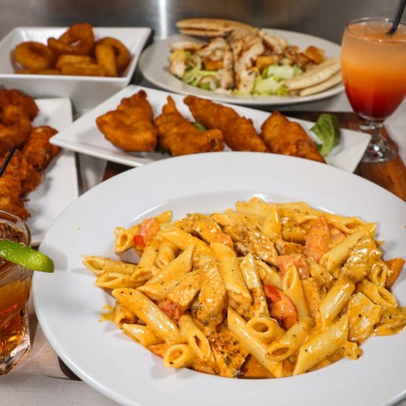 A table laden with American dishes including penne pasta, fried fish, onion rings, salad, and cocktails.